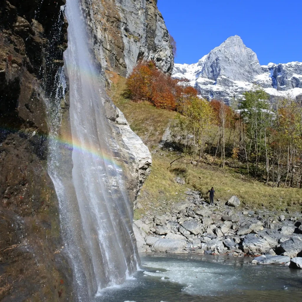 Un arc-en-ciel née dans un cascade avec les couleurs d'automne au 1er plan et les sommets déjà enneigés en arrière plan